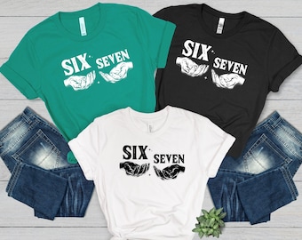 May include: Three t-shirts in teal, black, and white, each with the text "SIX SEVEN" above a graphic of two hands. The shirts are displayed with blue denim jeans. The shirts are made of a soft material.