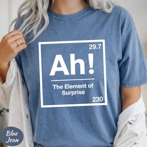 May include: Blue jean colored t-shirt with the text "Ah! The Element of Surprise" in a white box. The shirt has a crew neck and short sleeves. The text is in a bold, sans-serif font.
