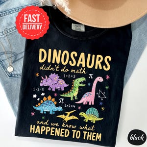 A black t-shirt with a humorous dinosaur-themed design, including a dinosaur with a math equation, and text that reads "DINOSAURS didn't do math and we know what happened to them".