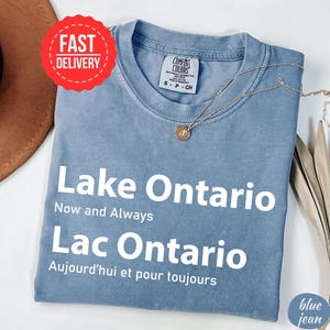 May include: A blue jean coloured t-shirt featuring the white text Lake Ontario Now and Always and Lac Ontario Aujourd'hui et pour toujours. The shirt has a Comfort Colors brand label at the neckline and is paired with a gold tone pendant necklace.