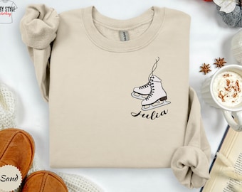 May include: Sand-colored sweatshirt with a crew neck. The design features a pair of white ice skates with black details and the name "Julia" in a cursive font. The sweatshirt is a neutral color.