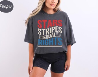 May include: Dark gray t-shirt with the text "STARS STRIPES EQUAL RIGHTS" in red, white, and blue. The shirt is worn with black shorts. The shirt has a vintage, worn look.