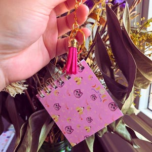 May include: A pink mini notepad with a spiral binding, featuring a repeating pattern of cat faces, stars, and houses. The notepad is attached to a gold keychain with a pink tassel.