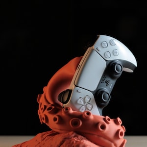 May include: A white and black video game controller rests in an orange octopus-shaped stand. The controller has multiple buttons and directional pads. The stand has a textured surface and suction cups.