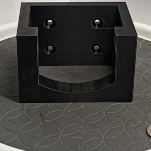 Op de afbeelding: Een zwart, 3D-geprint vierkant object met een gebogen uitsparing aan de onderkant en vier cirkelvormige gaten aan de voorkant. Het object staat op een gestructureerd oppervlak.