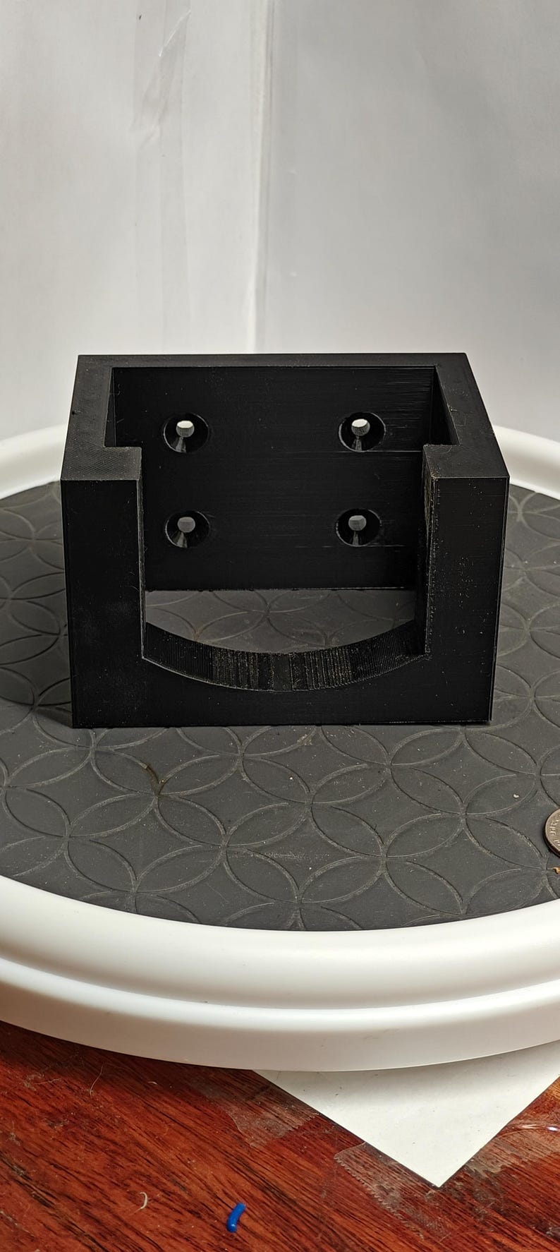 Op de afbeelding: Een zwart, 3D-geprint vierkant object met een gebogen uitsparing aan de onderkant en vier cirkelvormige gaten aan de voorkant. Het object staat op een gestructureerd oppervlak.