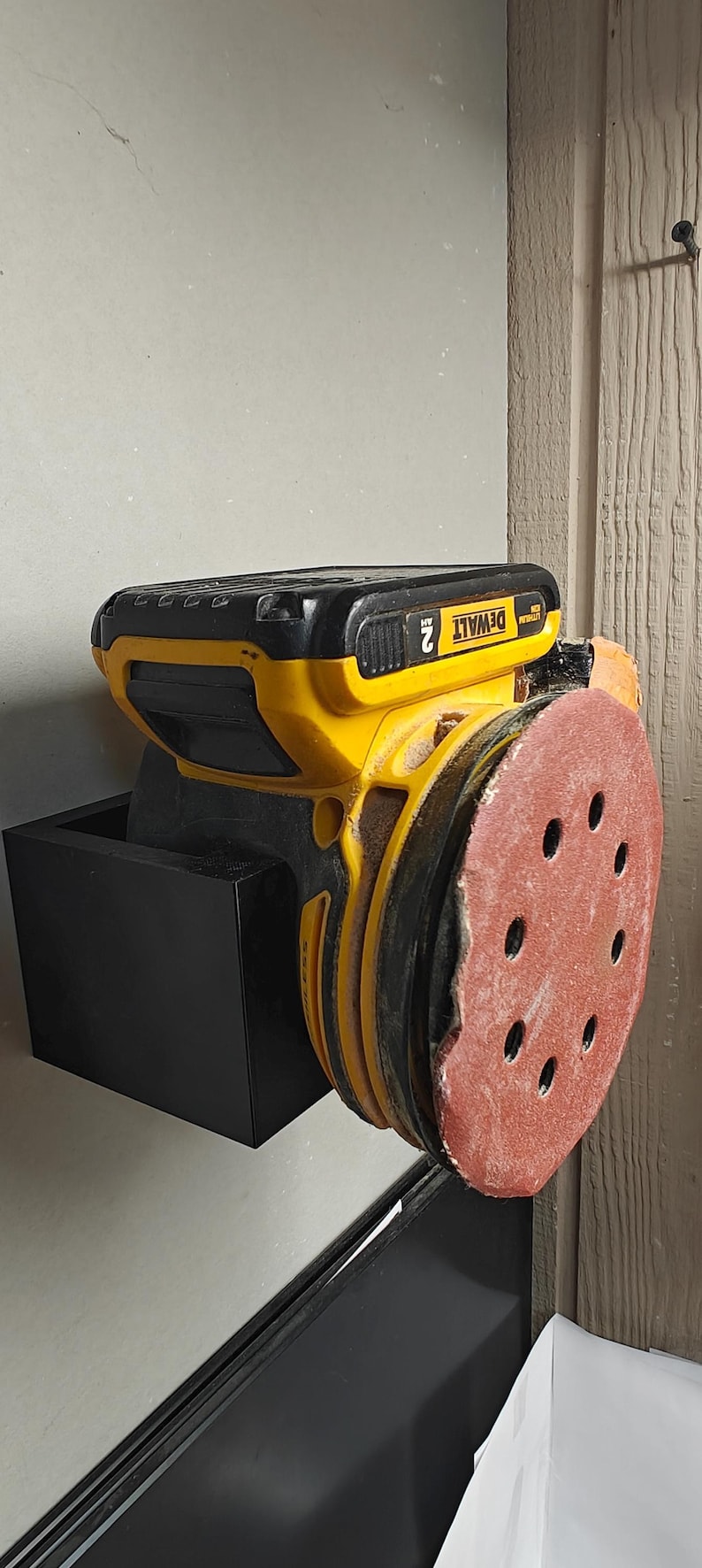Op de afbeelding: Een gele en zwarte DeWalt willekeurige orbitschuurmachine met een rode schuurpad. De schuurmachine is gemonteerd op een zwarte houder. De batterij geeft 2 AH aan.