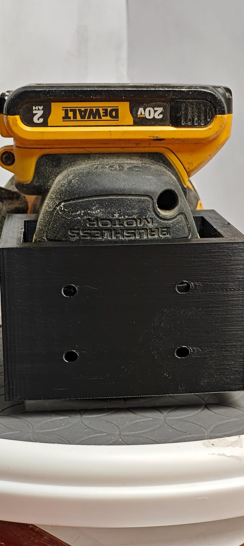 Op de afbeelding: Een gele en zwarte DeWalt 20V accuschuurmachine met een zwart opzetstuk. Op de schuurmachine staat de tekst "DEWALT" en "BRUSHLESS MOTOR". Het opzetstuk heeft vier gaten.