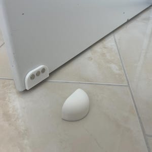 May include: A white cabinet door corner with a white door stop on a tiled floor. The door has a white corner piece with three silver rivets. A detached white door stop is on the floor.