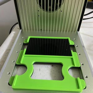 May include: An open, silver-colored machine with a black interior and a green frame holding a black rectangular object. The machine has a fan at the top and is on a white surface.