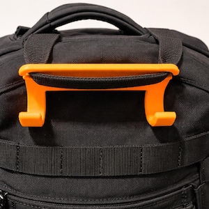 May include: Close-up of a black backpack with an orange handle attached to the top. The handle is a bright orange color and is designed to make carrying the backpack easier. The backpack has multiple straps and pockets.