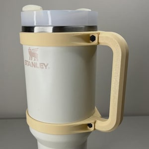 May include: A white Stanley travel mug with a light tan handle and accents. The mug has a silver rim and a clear lid. The Stanley logo is visible on the front of the mug. The mug is designed for travel and has a large handle.
