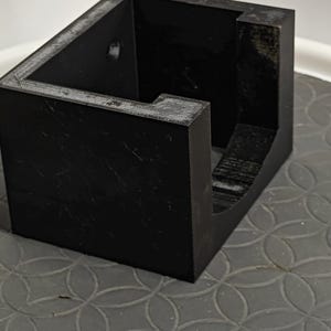 Op de afbeelding: Een zwarte, rechthoekige container met een open bovenkant en een uitgesneden gedeelte. Het object is gemaakt van een massief materiaal en staat op een oppervlak met een patroon. De container heeft een eenvoudig, geometrisch ontwerp.