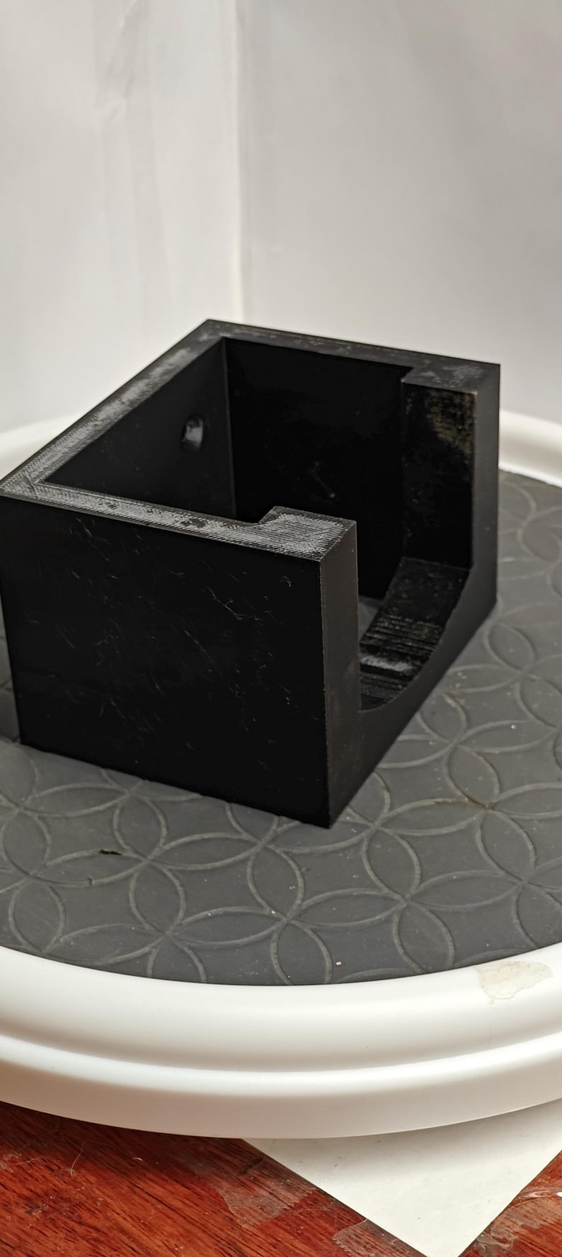 Op de afbeelding: Een zwarte, rechthoekige container met een open bovenkant en een uitgesneden gedeelte. Het object is gemaakt van een massief materiaal en staat op een oppervlak met een patroon. De container heeft een eenvoudig, geometrisch ontwerp.