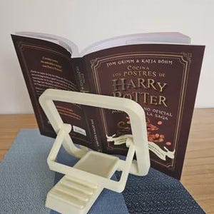 May include: A cream-colored book stand holds open a cookbook titled "Cocina Los Postres de Harry Potter." The book has a maroon cover with gold lettering. The stand is on a blue and gray patterned surface.