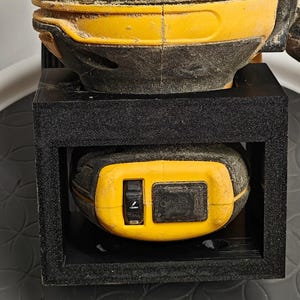 Op de afbeelding: Een gele en zwarte elektrische schuurmachine met een rode schuurpad. De schuurmachine zit in een zwarte houder. De schuurmachine staat op een zwart oppervlak met een munt op de voorgrond.