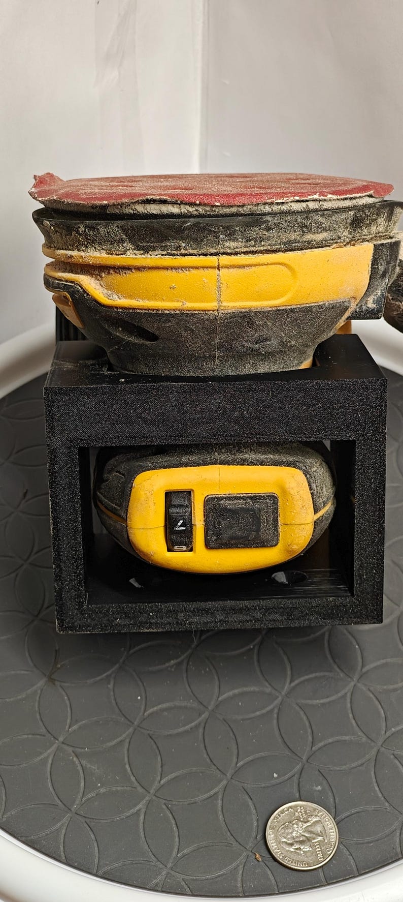 Op de afbeelding: Een gele en zwarte elektrische schuurmachine met een rode schuurpad. De schuurmachine zit in een zwarte houder. De schuurmachine staat op een zwart oppervlak met een munt op de voorgrond.
