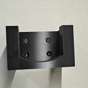 Op de afbeelding: Een zwarte, 3D-geprinte beugel met een U-vormig ontwerp. De beugel heeft vier schroefgaten en is gemonteerd met vier zilveren schroeven. Het item is waarschijnlijk een montagebeugel voor een elektronisch apparaat of iets dergelijks.