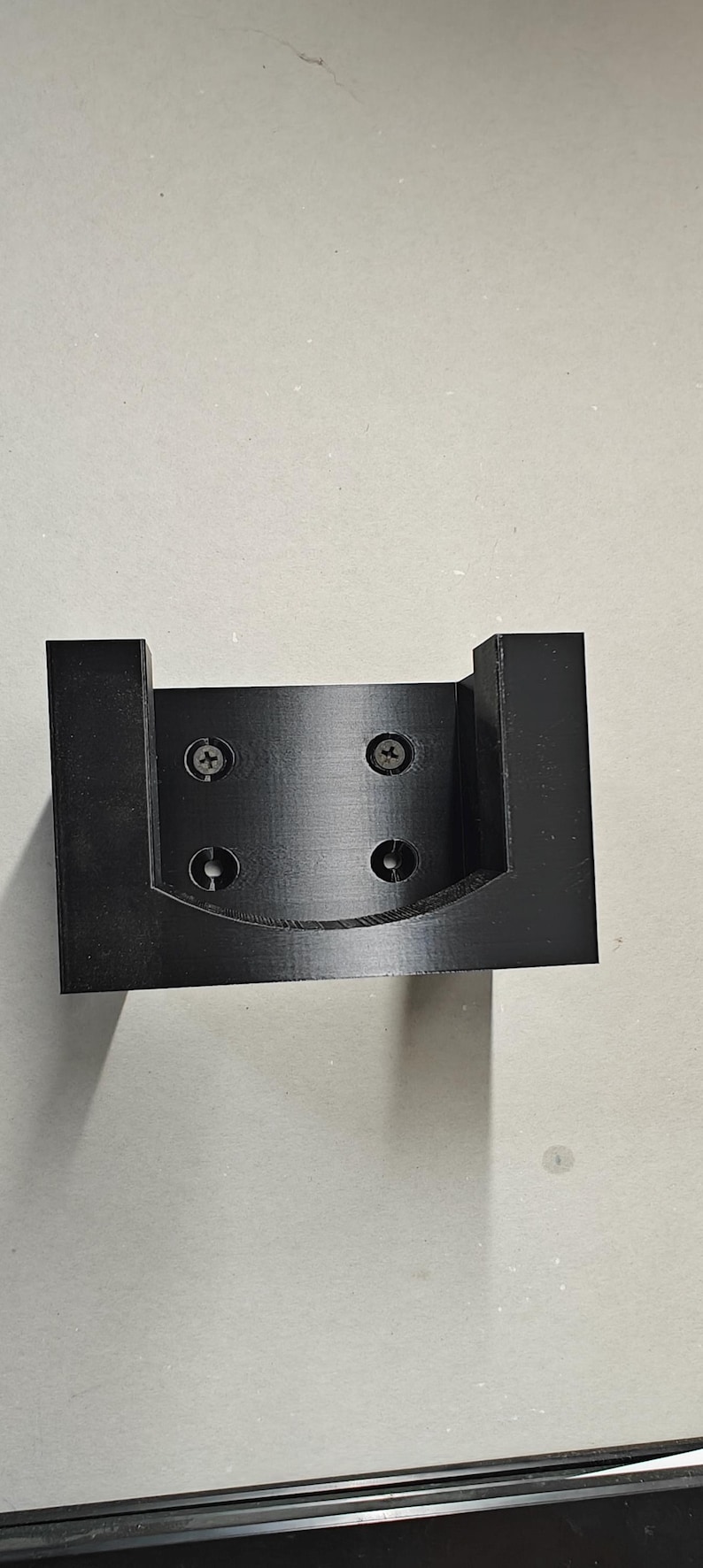 Op de afbeelding: Een zwarte, 3D-geprinte beugel met een U-vormig ontwerp. De beugel heeft vier schroefgaten en is gemonteerd met vier zilveren schroeven. Het item is waarschijnlijk een montagebeugel voor een elektronisch apparaat of iets dergelijks.
