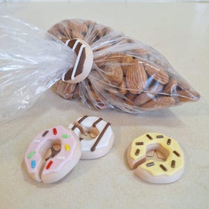 May include: A clear plastic bag filled with almonds is secured with a donut-shaped bag clip. Three additional donut-shaped clips in pink, white, and yellow with decorative sprinkles and icing are also visible.