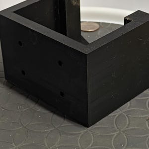 Op de afbeelding: Een zwart, L-vormig object met drie kleine gaten aan &eacute;&eacute;n kant. Het object is gemaakt van een massief materiaal en staat op een patroon. Het object lijkt een hoekbeugel of steun te zijn.