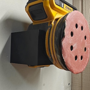 Op de afbeelding: Een gele en zwarte accuschuurmachine met een rode schuurpad. De schuurmachine is aan een muur bevestigd. De merknaam "DEWALT" is zichtbaar op de schuurmachine.