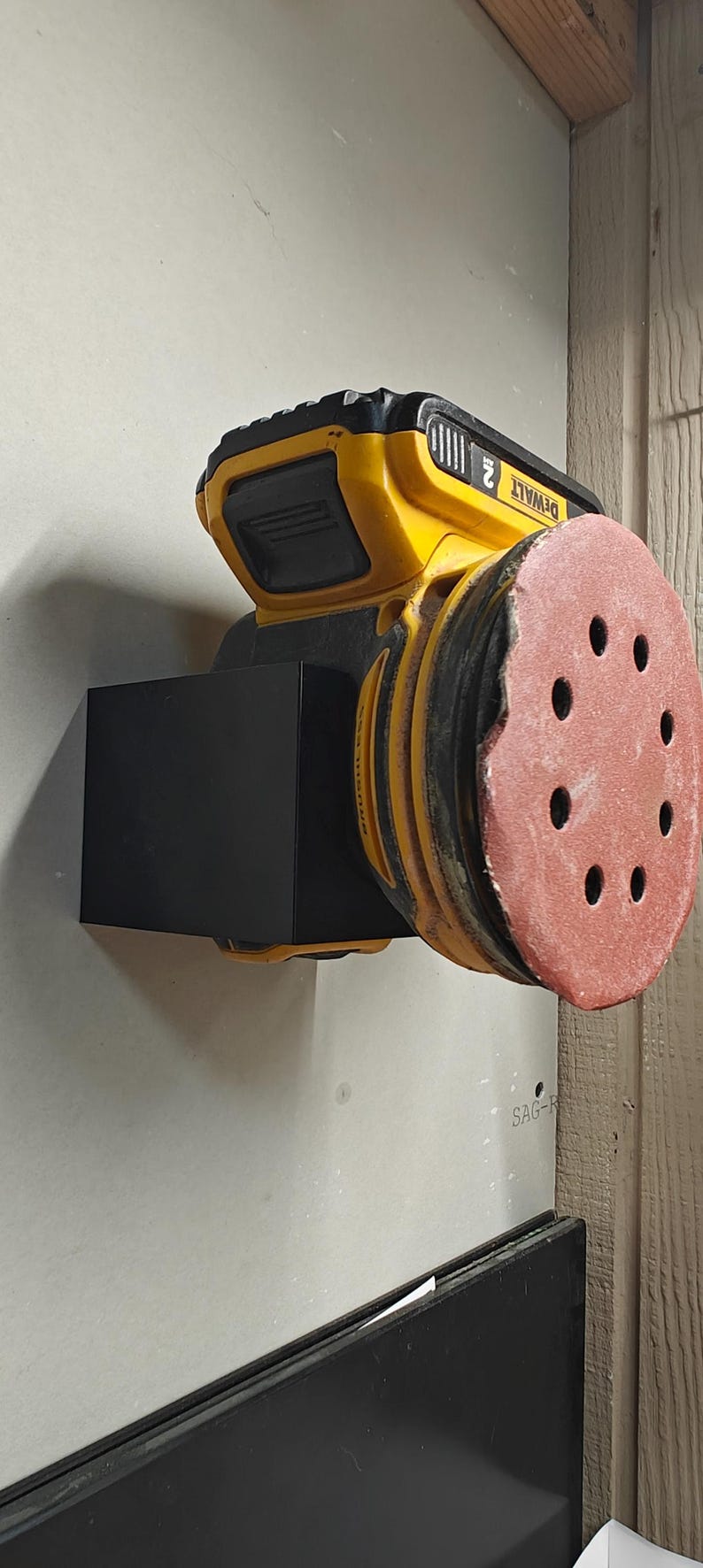 Op de afbeelding: Een gele en zwarte accuschuurmachine met een rode schuurpad. De schuurmachine is aan een muur bevestigd. De merknaam "DEWALT" is zichtbaar op de schuurmachine.
