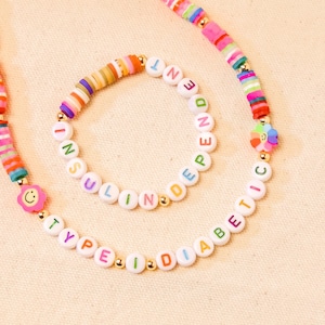 May include: Two colorful beaded necklaces with white beads spelling out "INSULINDEPENDENT" and "TYPE1DIABETIC". The necklaces are on a beige fabric background.