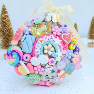 May include: A pink and blue Christmas ornament covered in glitter and decorated with candy, a rainbow, clouds, and the name "Gianna" with the word "Sweet" on a clear square.