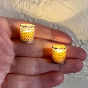 以下が含まれることがあります： 2つのミニチュアキャンドルホルダーと点灯したキャンドル。キャンドルホルダーは円筒形で、金色のダイヤモンド模様のケージデザインです。中のキャンドルは暖かく黄色い光を発しています。キャンドルホルダーは手のひらに収まるほど小さいです。