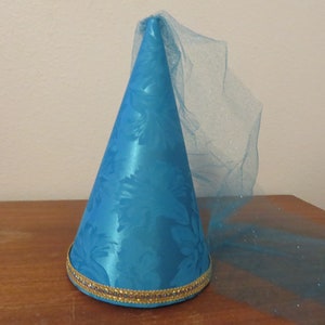 May include: A turquoise satin cone-shaped hat with a floral pattern and a gold trim at the base. A sheer, sparkly veil extends from the top. This festive hat is suitable for costume parties or themed events.