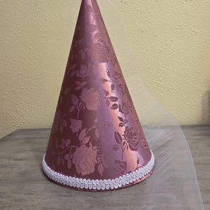 May include: A conical hat in a dusty rose color featuring a tonal floral jacquard pattern. The base of the cone is trimmed with a white braided decorative border, and a sheer white veil is attached to the top, draping down the side.