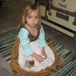 May include: A young girl wearing a costume dress with a brown bodice, light blue sleeves, and a white apron. The dress has a gold skirt and a heart-shaped patch. The girl is sitting on a striped rug.