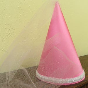 May include: A pink party hat with a white tulle veil. The cone-shaped hat is made of a shiny pink material and is trimmed with white decorative trim. The veil is attached to the hat and drapes down.