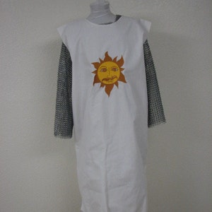 May include: A white sleeveless tunic featuring a central yellow and orange sun graphic with a human face. The tunic is layered over a long-sleeved metallic silver chainmail shirt. The garment is displayed on a mannequin against a plain wall.