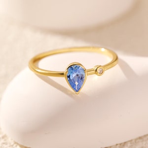 May include: A gold ring featuring a teardrop-shaped blue gemstone and a small round clear gemstone.