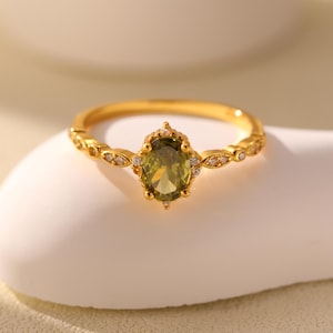 May include: A gold ring featuring a green oval gemstone surrounded by smaller clear gemstones. The ring has a delicate design with a band that is adorned with small, clear gemstones.
