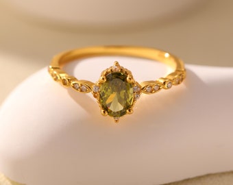 May include: A gold ring featuring a green oval gemstone surrounded by smaller clear gemstones. The ring has a delicate design with a band that is adorned with small, clear gemstones.