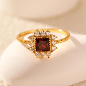 Könnte beinhalten: Ein goldener Ring mit einem roten quadratischen Edelstein, der von kleineren klaren Edelsteinen umgeben ist.