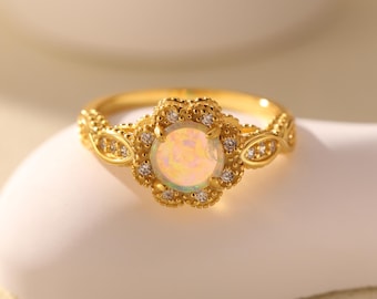 May include: A gold-colored ring with a central, iridescent, round gemstone. The ring features a floral design around the gemstone, with small, clear accent stones. The band has a detailed, textured pattern.