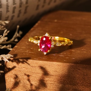 May include: A gold ring featuring a large oval ruby gemstone surrounded by smaller clear gemstones. The ring is displayed on a wooden surface.