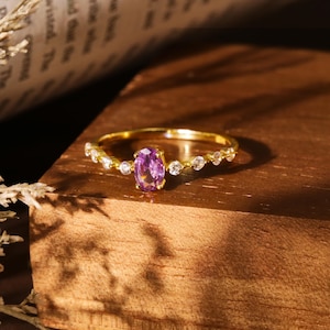 Puede incluir: Un anillo de oro con una piedra ovalada de color púrpura rodeada de piedras pequeñas transparentes.