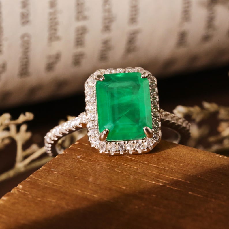 May include: A silver ring with a large emerald green gemstone surrounded by smaller diamonds. The ring is set on a wooden surface.