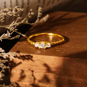 Könnte beinhalten: Ein goldfarbener Ring mit einem gedrehten Band und drei klaren, runden Edelsteinen. Der Ring liegt auf einer Holzoberfläche. Der Ring ist ein Schmuckstück.