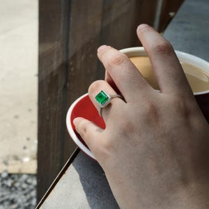 May include: A silver ring with a large emerald green gemstone surrounded by smaller clear gemstones.