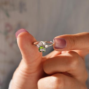 Puede incluir: Un anillo de plata con un diseño de huella de pata. El anillo presenta piedras claras y una piedra central iridiscente de color verde. El anillo es sostenido por una mano, destacando los delicados detalles de la joya.