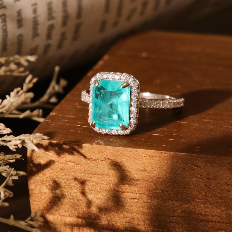 May include: A silver ring with a large, emerald-cut turquoise gemstone surrounded by a halo of smaller diamonds.