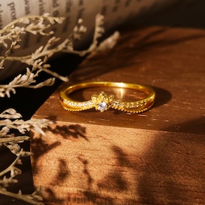 Può includere: Un delicato anello d'oro con una piccola pietra preziosa trasparente al centro. L'anello ha un design floreale ed è posto su una superficie di legno.
