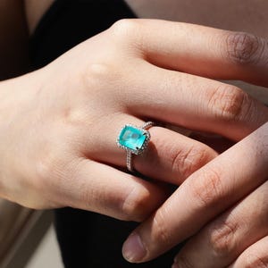 May include: A silver ring with a large, emerald-cut turquoise gemstone surrounded by a halo of smaller diamonds.