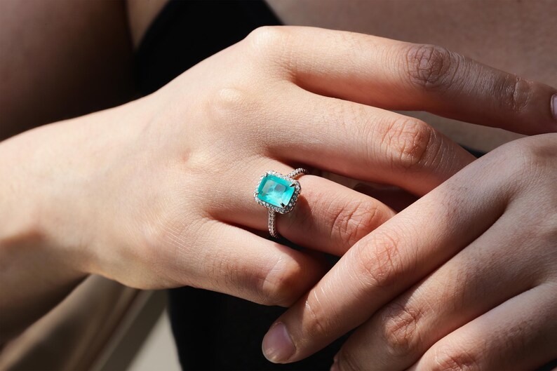 May include: A silver ring with a large, emerald-cut turquoise gemstone surrounded by a halo of smaller diamonds.